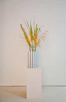 Silberne, skulpturale Vase mit wellenförmiger Oberfläche, arrangiert mit gelben Blüten und Ziergräsern, auf weißem Sockel vor minimalistischem Hintergrund. Szenografie in der Galerie Durchgang Basel. elena antoni, innenarchitektur, szenografie, kreative beratung, basel, schweiz, deutschland