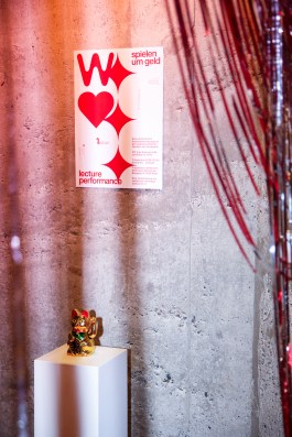 Plakat zur Lecture Performance „Spielen um Geld“ an roher Betonwand, inszeniert von Elena Antoni. Im Vordergrund eine goldene Winkekatze auf Sockel sowie silberne Lametta-Elemente – ein szenografisches Setting im Projektraum Wildwuchs, Basel. elena antoni, innenarchitektur, szenografie, kreative beratung, basel, schweiz, deutschland