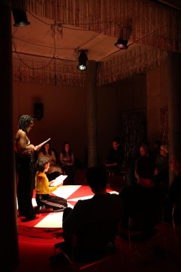 Szene der Lecture Performance Spielen um Geld im Projektraum Wildwuchs Basel: Zwei Performerinnen auf rot-weiss kariertem Podest unter Spotlight, umgeben von Publikum im Halbrund. Der Raum ist in warmes Licht getaucht, mit glitzernden Vorhängen und szenografischer Gestaltung von Elena Antoni. elena antoni, innenarchitektur, szenografie, kreative beratung, basel, schweiz, deutschland